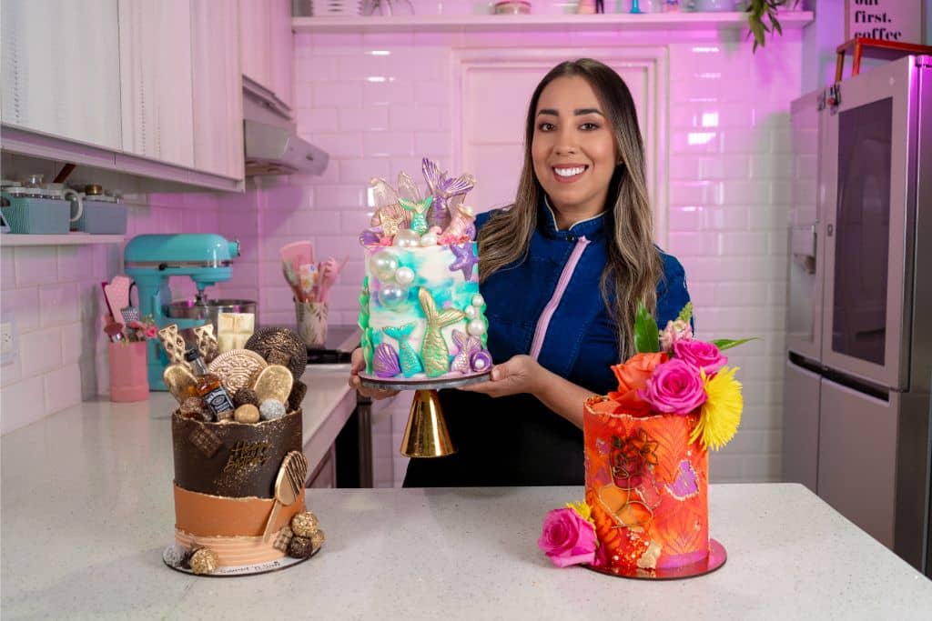 Bel García en su cocina rosada sosteniendo un pastel artístico de sirena, junto a un pastel de chocolate y otro floral